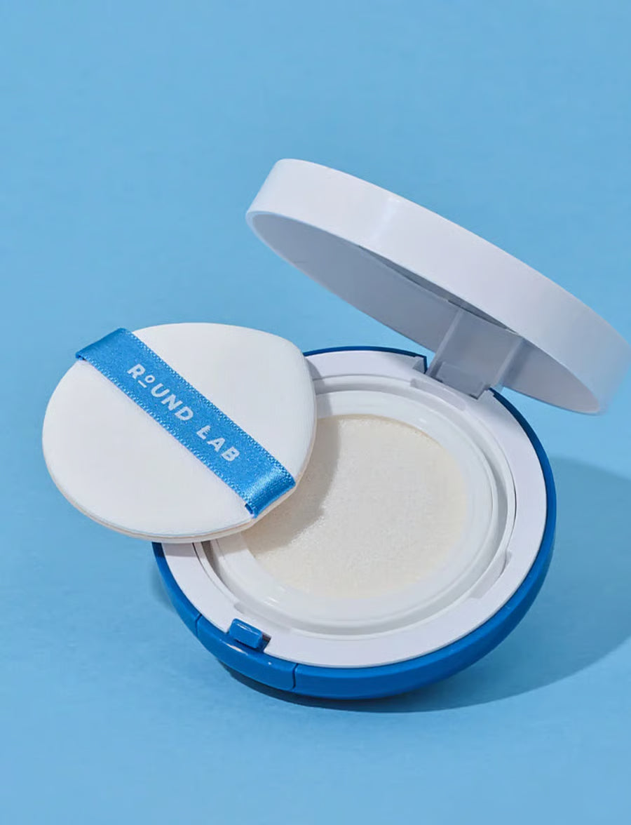 An open Round Lab cushion foundation compact on a light blue background, showing the white cushion foundation inside and the white puff applicator with a blue ribbon label.