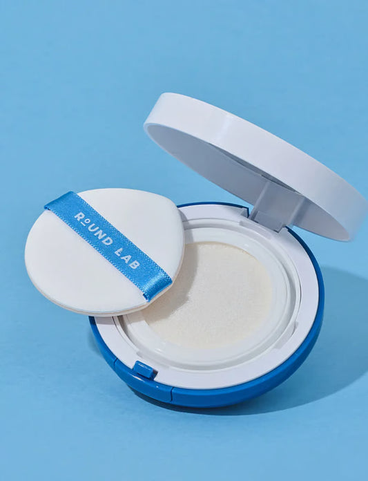An open Round Lab cushion foundation compact on a light blue background, showing the white cushion foundation inside and the white puff applicator with a blue ribbon label.